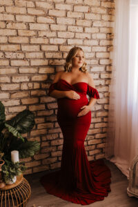 Mom posing in front of brick wall In red dress for maternity pictures