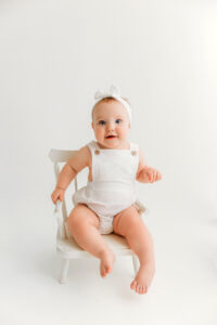 Baby girl in white outfit on white chair for infant photos in phoenix