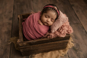 Baby posed in crate wrapped in pink wrap with headband for newborn photography session