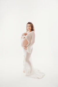 Hispanic mom posing Maternity portrait session in Phoenix on White backdrop with White maternity gown 