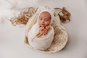 Baby swaddled in cream wrap in a heart shaped bowl for simple newborn session at phoenix photography studio holding a bear