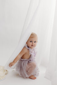 1 year old girl posing during a cake smash photoshoot in phoenix in purple outfit hiding behind curtain