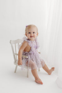 1 year old girl posing during a cake smash photoshoot in phoenix in purple outfit on white wood chair