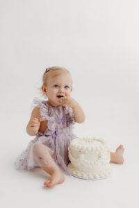 1 year old girl posing during a cake smash photoshoot in phoenix in purple outfit eating cake 