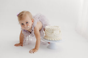 1 year old girl posing during a cake smash photoshoot in phoenix in purple outfit climbing over cake