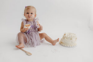 1 year old girl posing during a cake smash photoshoot in phoenix in purple outfit eating cake